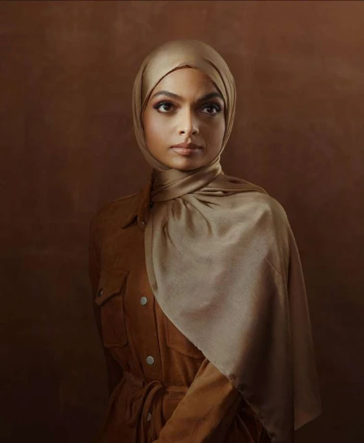 Portrait of a woman with dark hair wearing a brown hijab and matching jacket against a blended brown backdrop in a photo studio.